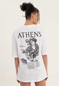 Vrouw met gevlochten haar die een wit t-shirt draagt met de tekst "ATHENS", een klassieke buste omwikkeld door een draak, en kleine architectonische schetsen op de achterkant.