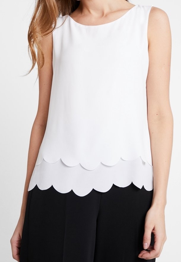 Blouse blanche sans manches avec bords en couches en forme de coquille portée avec un pantalon noir sur une personne aux cheveux longs.