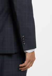 Dunkle kariertes Sakko mit glatter Textur, das einen Vier-Knopf-Manschettenverschluss und einen weißen Hemdmanschettenrand zeigt, der unter dem Ärmel hervorlugt.