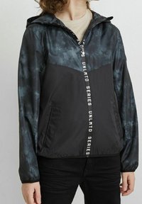 Chubasquero con capucha en patrón negro y gris, con cierre de cremallera y texto vertical en blanco "UNLMTD SERIES" en el frente.