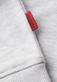 Tessuto grigio dalla texture morbida, con un'etichetta rossa visibile che recita "SUPERDRY". Design semplice con linee pulite e dettagli sottili.
