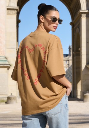 Femme aux cheveux foncés en chignon porte un T-shirt marron oversize avec un texte rouge en forme de cœur dans le dos et un jean bleu clair, debout en plein air.