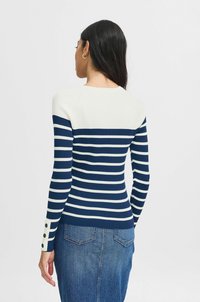 Pull rayé bleu marine et blanc avec une texture côtelée, doté de longues manches et de boutons décoratifs aux poignets, associé à une jupe en denim.