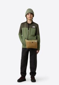 Roheline fliisjakk luku ja esitaskutega, mustad püksid ja pruun risti kantav kott. Triibuline müts täiendab outfiti.