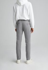 Graue Baumwollchinos mit Slim-Fit, ausgestattet mit einem Knopf- und Reißverschluss, Gesäßtaschen und dezenten Nähdetails. Weiße Sneakers sind sichtbar.