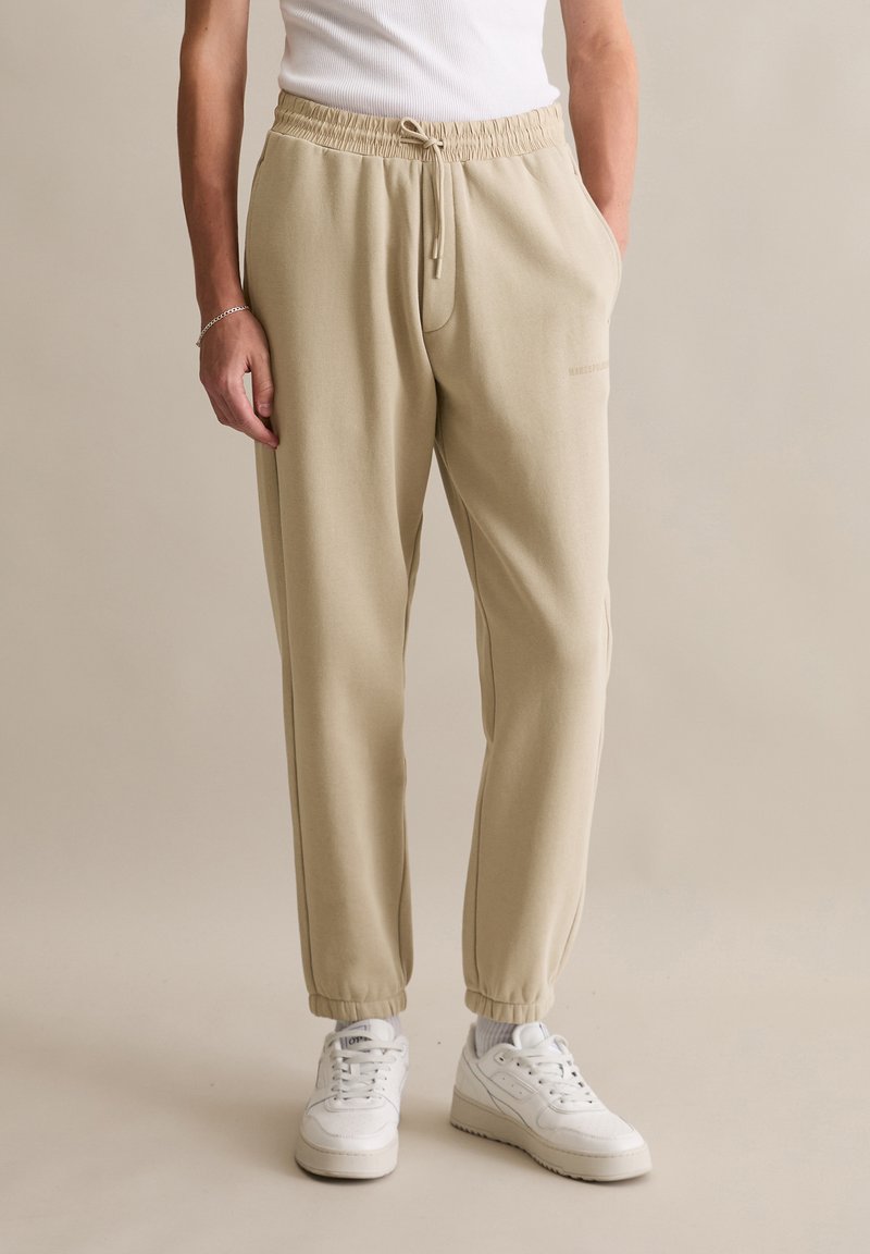 Beige joggingbroek met een elastische tailleband en trekkoord, voorzien van zijzakken en taps toelopende boorden. Gecombineerd met witte sneakers.