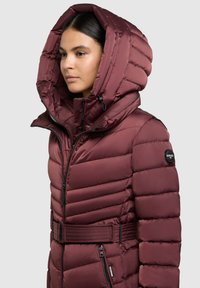 Bordo puhovka z veliko kapuco, prešitim vzorcem, zadrgo spredaj, stranskimi žepi in pasom. Na rokavu je vidna logotip.