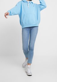 Niebieska bawełniana bluza z kapturem, kieszenią z przodu i sznurkiem przy kapturze, zestawiona z jasnoniebieskimi obcisłymi dżinsami i białymi wysokimi trampkami.