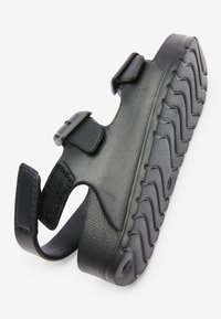 Sort sandal med en tekstureret gummisål, justerbare stropper og åben hældesign. Har udtalt mønster for greb og sikker pasform.