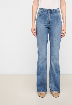 Kvinde iført lyseblå højttaljede jeans med lige ben og beige balletsko, stående mod en ensfarvet hvid og beige baggrund.