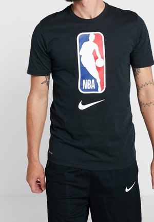 Mann trägt ein schwarzes Nike Dri-FIT T-Shirt mit NBA-Logo und schwarze Nike-Shorts, steht mit sichtbaren Arm-Tattoos.
