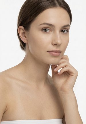 Jonge vrouw met blote schouders die kleine gouden oorbellen in hartvorm draagt, haar kin steunend op haar hand, met een neutrale gezichtsuitdrukking.