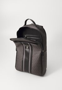 Prugasti ruksak tamnosive boje pravokutnog oblika s crnim okomitim prugama i metalnim V emblemom, djelomično otvoren kako bi se vidjela unutarnja zip zatvorena torbica.