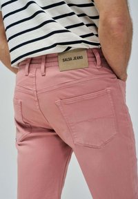 Svetlo roza Salsa jeans hlače z gladko teksturo, z usnjenim etiketom na zadnjem pasu in dvema zadnjima žepoma. Črtasta srajca je vidna zgoraj.