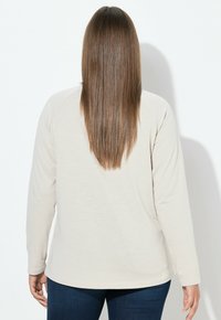 Ljust beige långärmad topp med avslappnad passform och raglanärmar, kombinerad med mörkblå jeans. Slätt tyg och rundad nederdel.