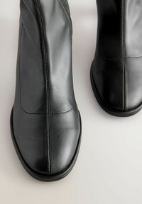 Stivali alla caviglia in pelle nera con una texture liscia, caratterizzati da cuciture verticali e un design a punta arrotondata. La suola è piatta e semplice.