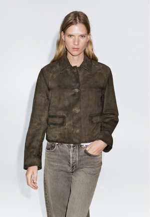 Giacca corta in suede marrone con colletto, due tasche frontali e chiusura con bottoni. Indossata sopra jeans grigi chiari. Superficie semplice e testurizzata.