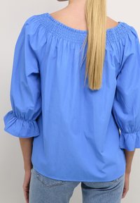 Blouse bleue avec encolure smockée, manches bouffantes trois-quarts et coupe décontractée. Fabriquée en tissu léger à texture lisse.