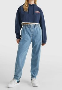 Sudadera navy recortada con interior blanco, que presenta un logo rojo; jeans de talle alto de color azul claro con cintura fruncida; zapatillas blancas.