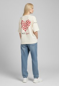 T-shirt crème à manches courtes avec un grand motif de cœur rouge et le texte « SWEET heart » dans le dos, associé à un jean bleu clair.