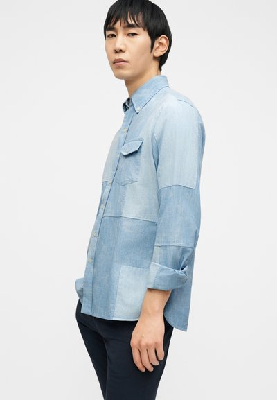 Νέος άνδρας που φοράει ένα ανοιχτό μπλε πουκάμισο από denim με μπαλώματα, με τα μανίκια γυρισμένα, στέκεται πλάγια μπροστά σε λευκό φόντο.