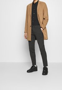Cappotto lungo in lana color tabacco, camicia nera con bottoni, pantaloni scuri a righe sottili e sneaker nere. Design minimalista con colori a contrasto.