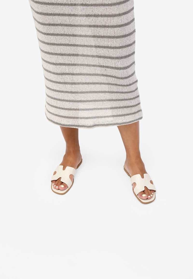 Personne portant une jupe beige et grise à rayures et des sandales blanches ouvertes à l'avant, debout sur un sol blanc.