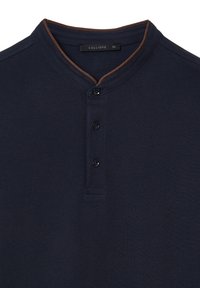 Polo blu navy con tessuto testurizzato, dotato di un accento marrone sul colletto, tre bottoni neri e un'etichetta che recita "CALLIOPE XL."