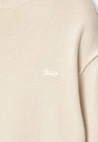 Pull tricoté couleur crème avec col côtelé et petit logo blanc brodé sur le côté droit de la poitrine.