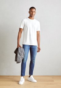 White short-sleeve t-shirt, blue skinny jeans, holding a gray jacket. White sneakers. Simple design, smooth texture on a wooden floor.