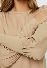 Beige Langarmtop mit runder Ausschnittlinie, das mit Rüschendetails an den Ärmeln und einer glatten, weichen Stofftextur ausgestattet ist.
