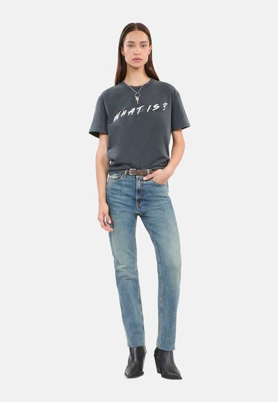 T-shirt en coton gris arborant la phrase « QUE EST-CE ? » en lettres blanches en gras, associé à un jean droit bleu clair et des bottines noires.