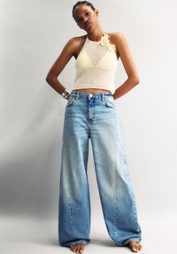 Szydełkowy top na halter w kolorze kremowym z kwiatowymi akcentami, w zestawieniu z szerokimi, wyblakłymi niebieskimi dżinsami. Prosty fason i luźny krój.