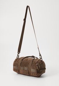Brown shearling crossbody bag with a cylindrical shape, featuring dual handles, adjustable strap, and flat webbing accents.