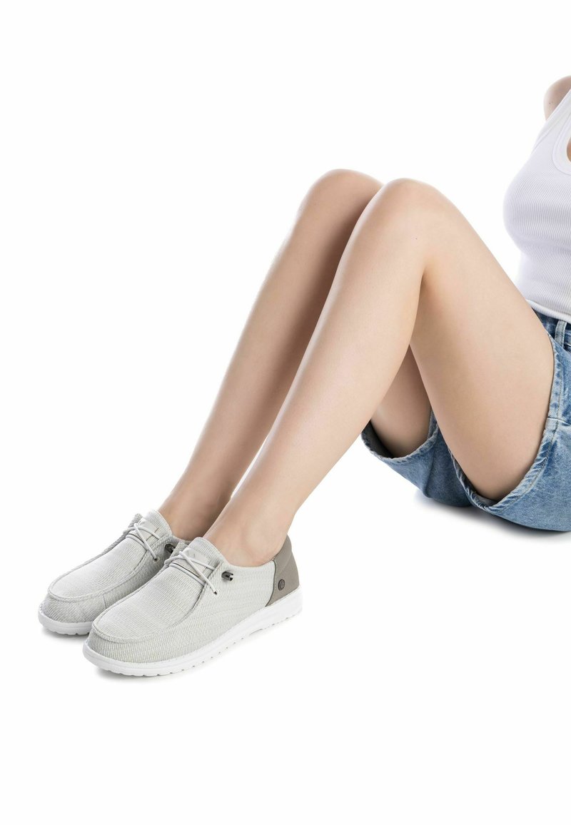 Zapatos textil de color gris claro con suela blanca, diseño de punta redonda, acentos en contraste en gris claro y beige, y un detalle decorativo de cordones en la parte superior.