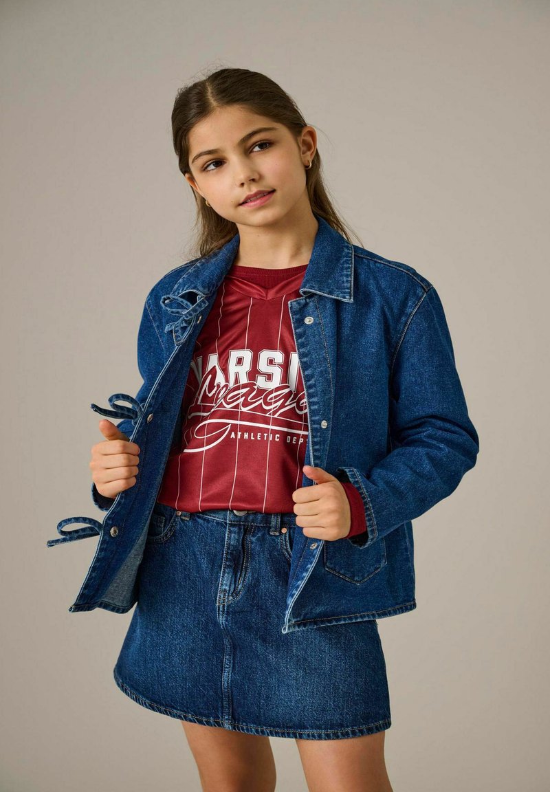 Denimu jaka ar āķa un cilpas aizdari, saskaņota ar sarkanu un baltu svītrotu kreklu un denimu svārkiem. Liegs tekstūra un ikdienišķs piegriezums.