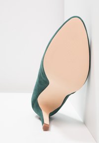 Chaussure à talon haut en daim vert à bout pointu reposant sur le côté, montrant la semelle beige lisse et le talon fin avec embout en caoutchouc.