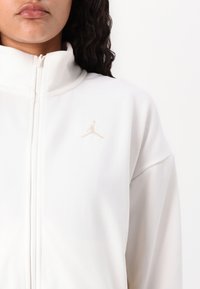 Hvid zip-up jakke med højt krave, prydet med et lyst beige broderet logo på brystet, lavet af glat stof.