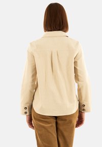 Chaqueta de pana beige con mangas largas, un collar con punta y puños abotonados. Presenta un plisado en la parte posterior para un detalle adicional.