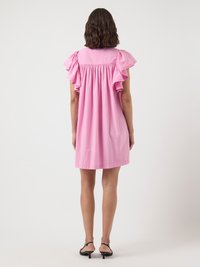 Sieviete, tērpusies brīvā rozā kleitā ar volāniņiem īsām piedurknēm, stāv pretējā virzienā, apavi ar melniem papēžiem uz baltas fona.