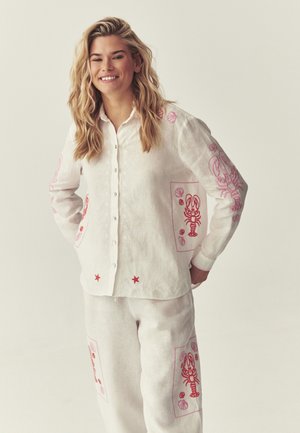 Lächelnde Frau mit blonden, welligen Haaren, die ein weißes Hemd mit Knöpfen und eine Hose mit roten Hummer- und Muschelmotiven trägt.