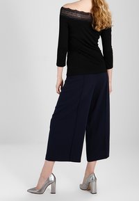 Svart off-shoulder topp med spetsdetaljer, tillsammans med marinblå vida byxor och silverfärgade högklackade skor. Slät textur, elegant silhuett.