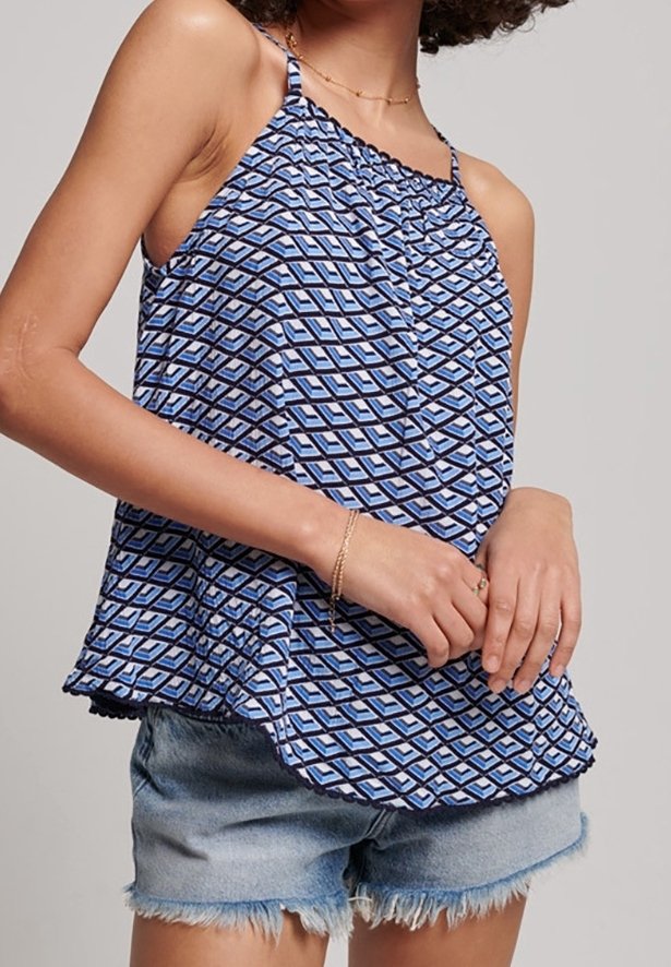 Femme portant un débardeur bleu à motif géométrique et un short en jean clair effiloché, avec des bijoux en or délicats au cou et au poignet.