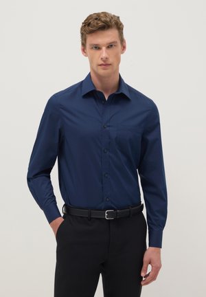 Mann mit lockigem Haar, trägt ein marineblaues langärmliges Hemd mit Knöpfen und schwarze Hose, steht mit einer Hand in der Tasche vor einem einfarbigen Hintergrund.