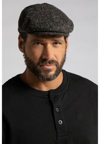 Gorra plana hecha de un tejido oscuro y estampado, con un diseño de espiga. Usada con una camisa de cuello abotonado negra. Fondo neutro.