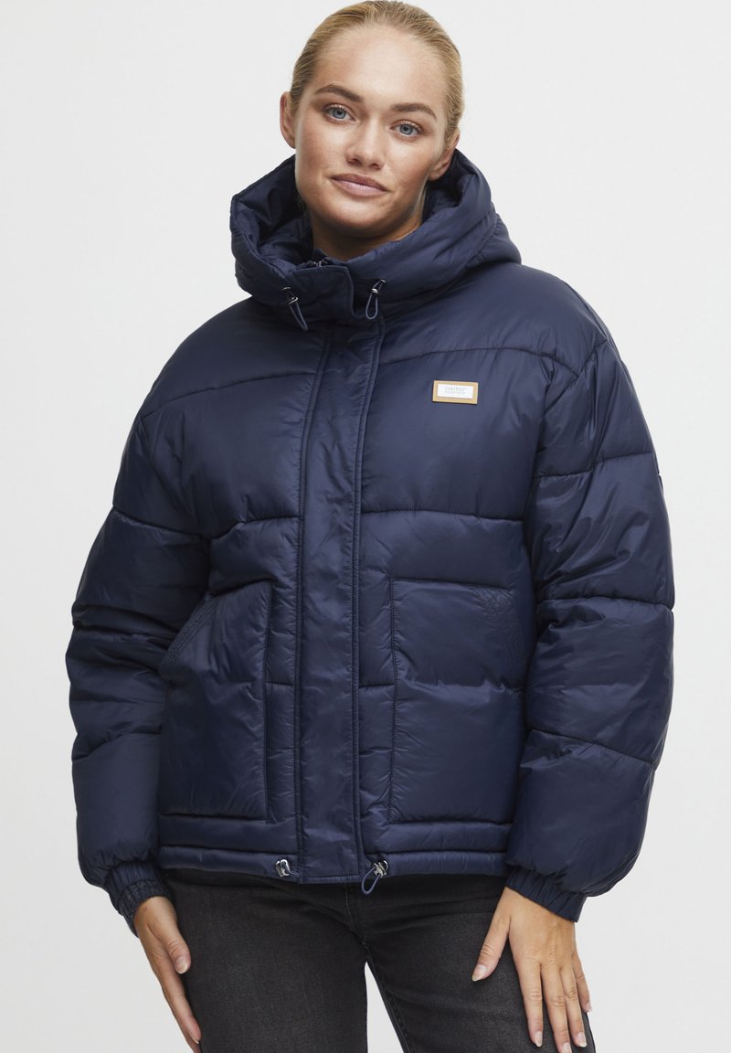 Giacca imbottita blu navy con colletto alto, chiusura a zip frontale e coulisse regolabile. Presenta una texture trapuntata e due tasche anteriori.