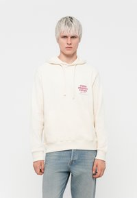 Cream-colored hoodie with a front pocket and a drawstring hood. Features red text stating "STUDIO ORIGINALS EDITION" on the chest.