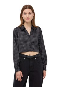 Blusa nera a cropped con finitura in satin, pois bianchi, maniche lunghe e colletto classico, abbinata a pantaloni neri a vita alta.