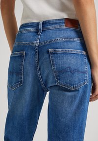 Close-up van een persoon die blauwe spijkerbroeken draagt met een gestikt golfpatroon op de achterzakken en een bruin lederen merkembleem op de tailleband.