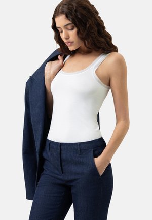 Frau mit welligem braunem Haar, die ein weißes ärmelloses Oberteil mit Spitzenbesatz, marineblaue, maßgeschneiderte Hosen trägt und einen passenden marineblauen Blazer hält.